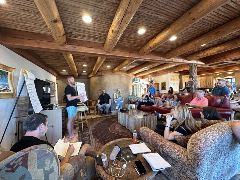 A presenter speaks to a seated group taking notes at a mastermind session in a wood-beamed room.