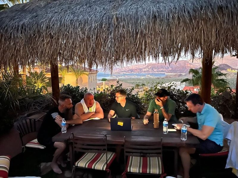 A group of men working on a laptop at a table under a thatched palapa overlooking a lake at sunset.